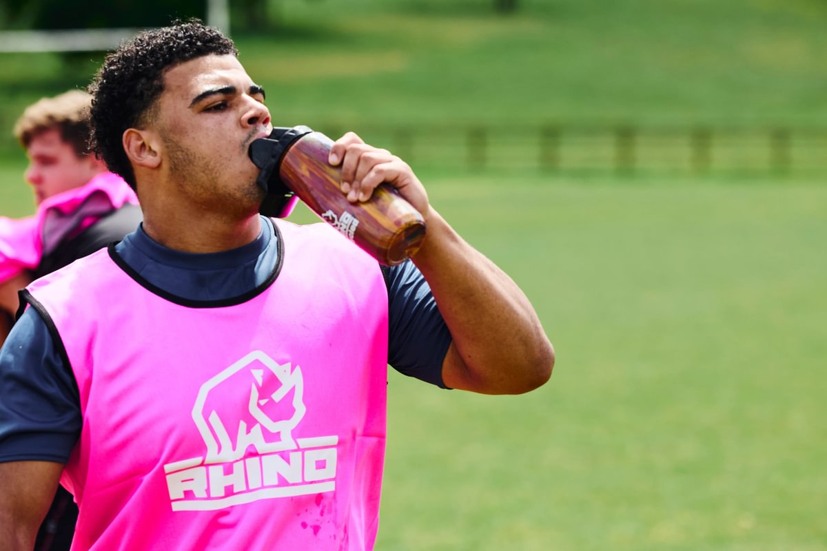 Person in a pink 'Rhino' bib drinking from a water bottle at rugby training on a pitch
