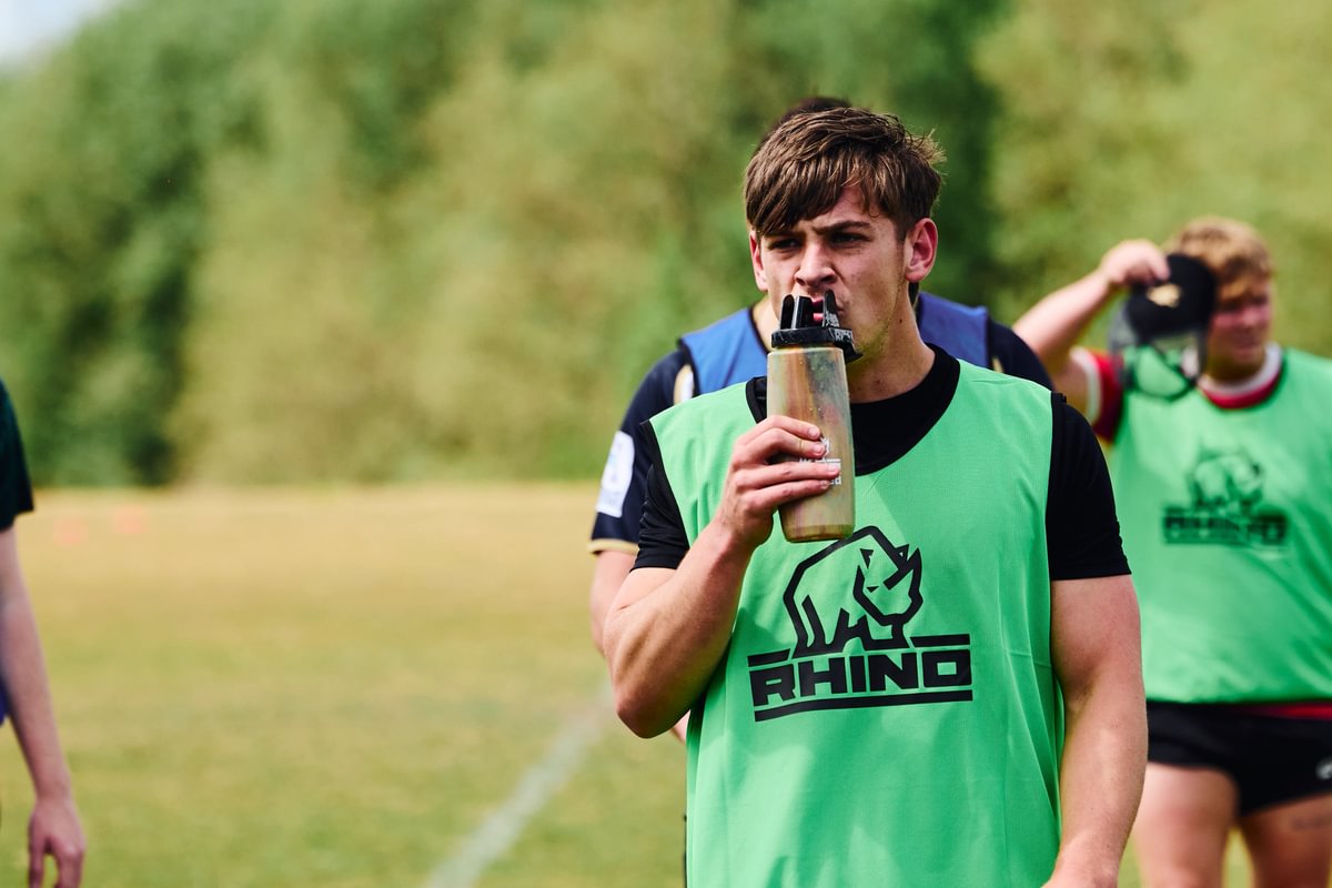 Person in a green training bib with a 'Rhino' logo drinking from a bottle on a rugby pitch