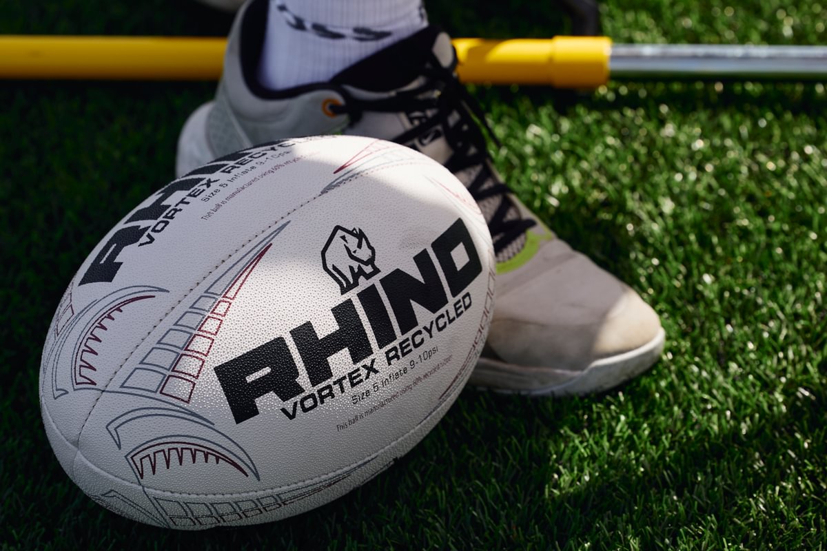 Rhino branded rugby ball on grass with a foot wearing a white shoe in the background