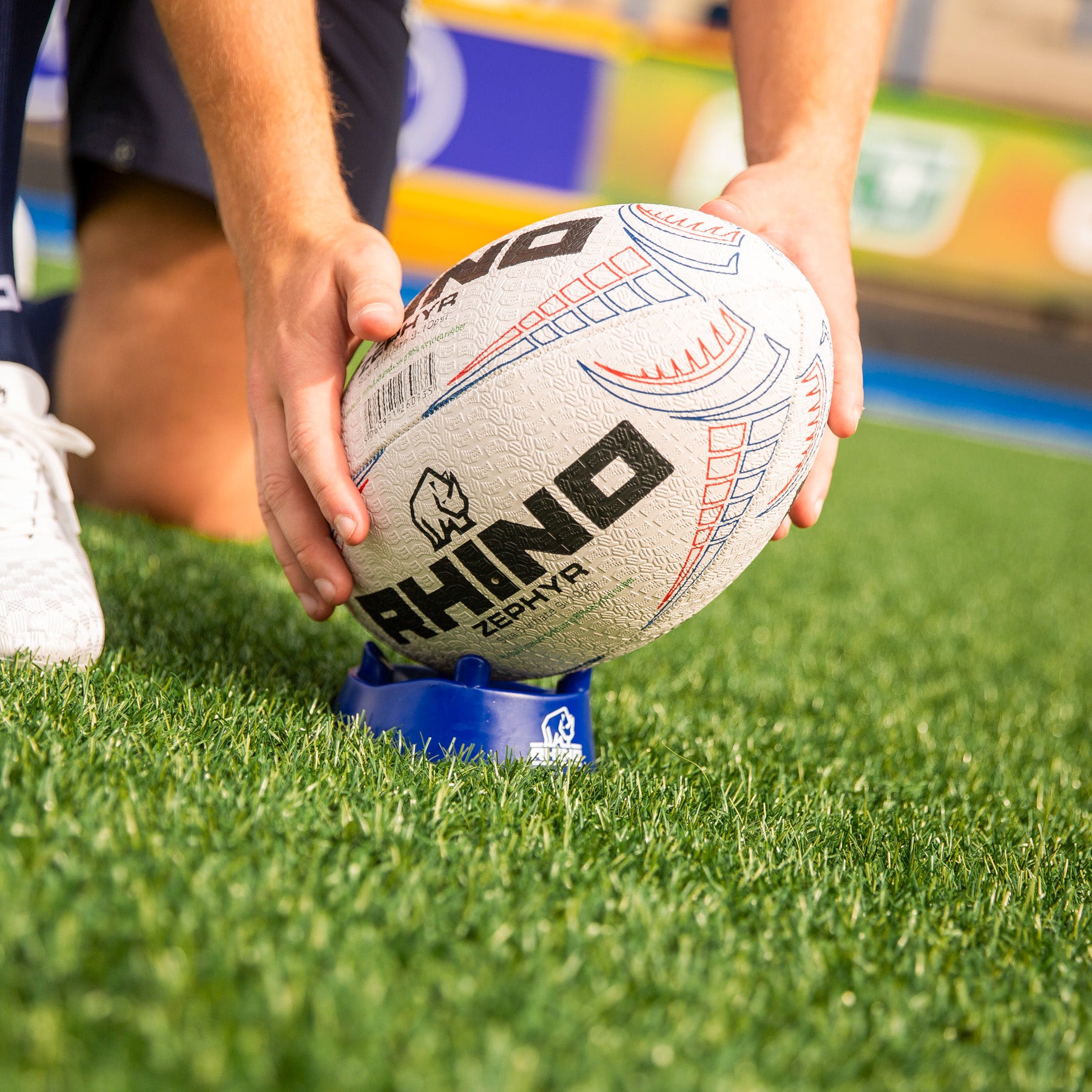 Person placing a rugby ball on a kicking tee on grass field with a Rhino brand logo.