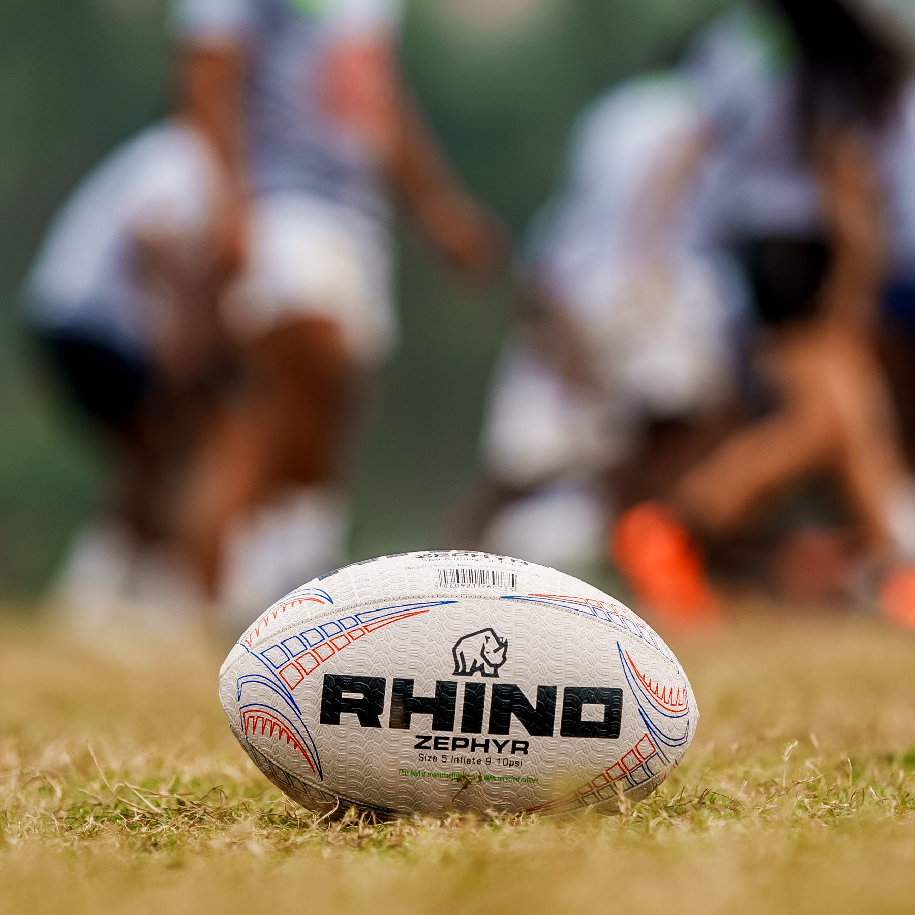 Rhino Zephyr rugby ball on grass with blurred players in the background